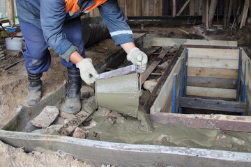 Working man in overalls pours concrete from a bucket on the floor around the garage observation pit, independent village indoor industrial construction building