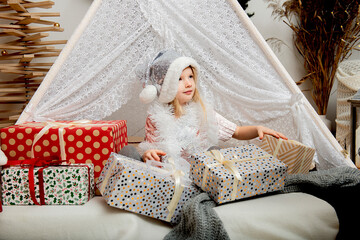 Portrait of a cute little girl child wearing a silver color Christmas hat sitting between gift boxes in a decorated house.Merry Christmas and Happy Holidays!