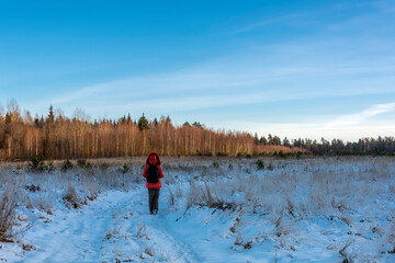 A woman with a backpack walks in the field along a snow-covered dirt road against the background of a forest winter landscape.
