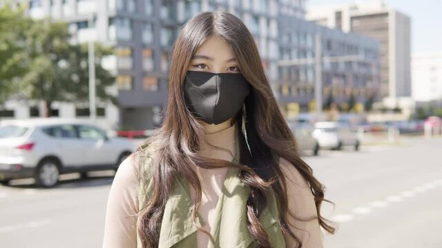 A young Asian woman in a face mask looks at the camera in a street in an urban area - cars drive on the busy road in the blurry background