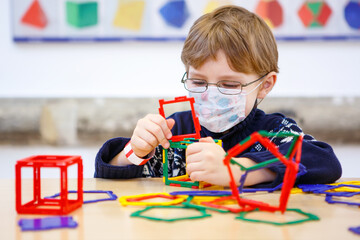 Little child with medical mask playing with lots of colorful plastic blocks kit in preschool...