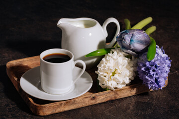 Hot coffee in white cup, milk and flowers hyacinth on wooden tray. Coffee, milk and flowers in low key on the dark background. 8 March concept