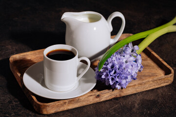 Hot coffee in white cup, milk and flowers blue hyacinth on wooden tray. Coffee, milk and flowers in low key on the dark background. 8 March concept