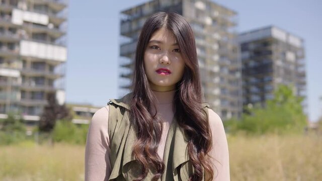 A young Asian woman shows a thumb up to the camera and nods with a smile - an apartment building complex in the blurry background