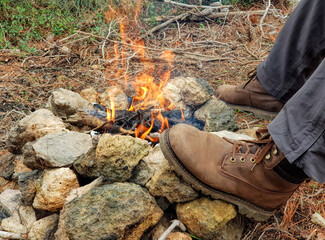 Traveler by the fire in the forest. Her feet are in rough hiking boots. Bonfire at stones. Burning wood and coals fire. Glow flame