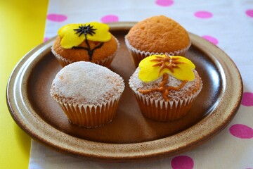 Plate of muffins decorated with powdered sugar and yellow icing in the shape of yellow flowers on a white tablecloth with pink polka dots on a yellow table