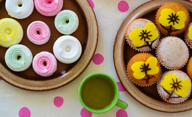 Overhead view of a plate of colored meringue cookies and another plate of muffins decorated with flower-shaped icing and a green cup of tea on a white tablecloth with pink polka dots