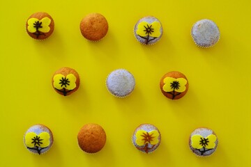Rows of muffins decorated with powdered sugar and yellow flower shaped icing on a yellow background