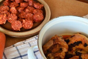 Pot with several roasted chicken pieces next to a plate of sliced ​​chorizo ​​on a white blue checkered cloth on a wooden table