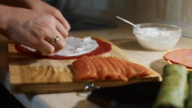 Thin tasty colorful pancakes. Close-up of woman's hand laying salmon slices as filling on red pancake anointed with creamy cheese on a wooden board. Rolling the pancake. 4K video.
