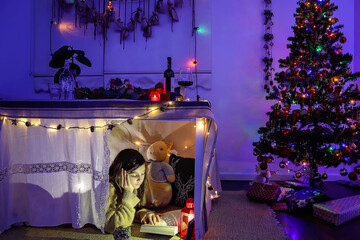 A girl reading a book in the fort next to the Christmas tree.