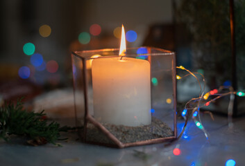 A burning candle in a glass holder in an atmospheric cozy atmosphere at home at night by the light of a garland. Dark light.  Holiday, Christmas and new year concept.
