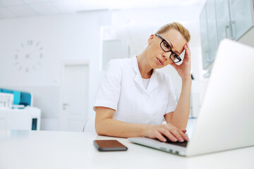 Serious dedicated gorgeous blond lab assistant with eyeglasses and in white sterile uniform sitting in laboratory, holding her head and entering data for research.