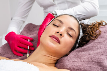Attractive young woman is getting a rejuvenating facial injections. She is sitting calmly at clinic. The expert beautician is filling female wrinkles by hyaluronic acid.