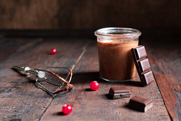 A glass jar with chocolate mousse, a whisk, pieces of dark chocolate and red currant berries on the...