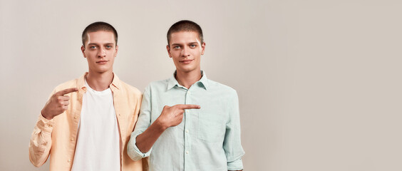 Two young twin brothers in casual wear pointing to the right and looking at camera while standing...