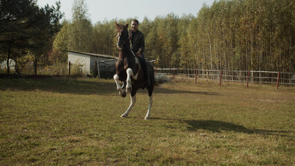 A young student of an equestrian riding school trains in an open-air paddock, riding a spotted...