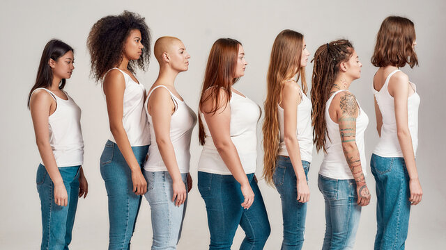 Group of beautiful diverse young women wearing white shirt and denim jeans looking aside while posing, standing isolated over grey background