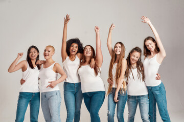 Group of beautiful diverse young women wearing white shirt and denim jeans having fun, looking cheerful while posing together isolated over grey background
