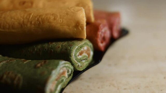 Thin tasty colorful pancakes. Close-up view of red, yellow and green cutaway rolled pancakes with cream cheese and salmon filling on a dish. 4K video.