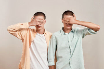 Portrait of two twin brothers covering eyes with hands while posing together isolated over beige background