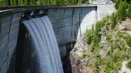 Water overflowing above an arched concrete dam built between two rocky mountain ridges covered with...