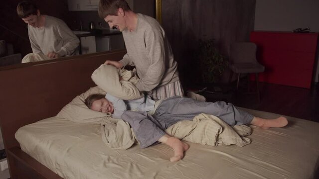 A young husband frolics with his wife in bed, they fight with pillows, he gently smacks her buttocks with his palm