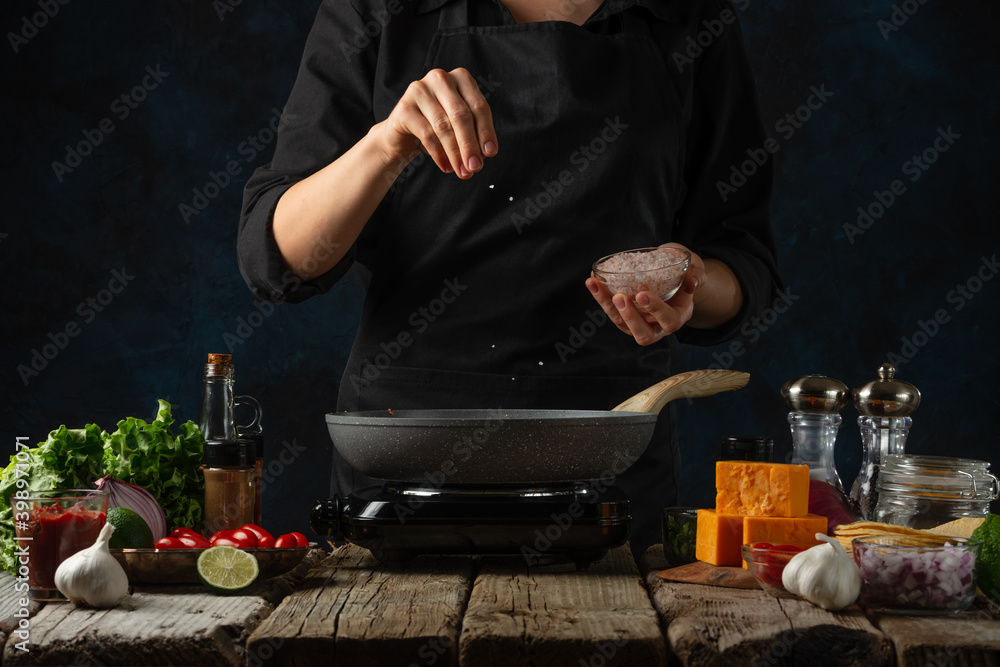 Wall mural Professional chef in black uniform pours sea salt into frying pan with meat filling. Backstage of cooking traditional mexican tacos on dark blue background. Frozen motion. Cookbook illustration. - Wall murals