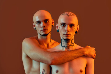 Portrait of young half naked twin brothers with tattoos and piercings looking at camera, posing together, standing isolated over orange background