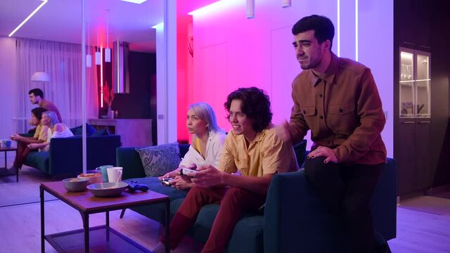 Smiling friends playing video games at home. The guy support and gives advices to his male friend. They celebrate victory and laughing.
