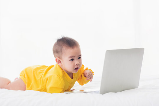 Asian little baby girl wearing a yellow dress typing hands on laptop computer keyboard and looking at monitor on the white bed. Little IT guy