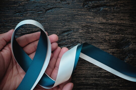 Top view of hand holding white and teal ribbon on dark wooden background with copy space. Cervical cancer awareness month, care and hope concept.