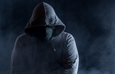A hooded man with a face mask waits in the dark