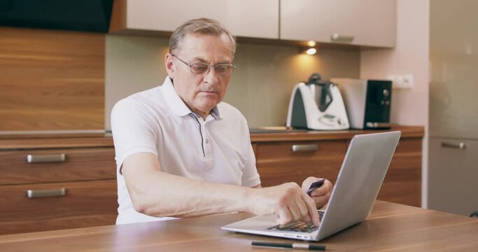 Warm toned portrait of modern senior man shopping online or paying taxes holding credit card while using laptop in kitchen at home.