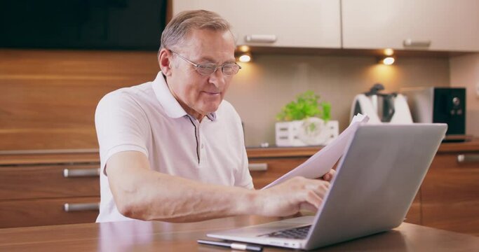 Happy elderly man sitting on chair in kitchen and buying with laptop use online website, voucher and bank credit card booking hotel plan vacation, old man learning payment system in internet.