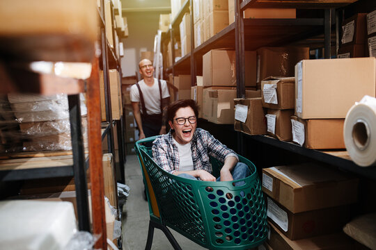 Warehouse Workers Having Fun During Working Shift, Man Pushing Cart With Woman, Accelerating And Letting Her Go With Some Speed. In A Passage Between Rows Of Boxes. Both Laughing.