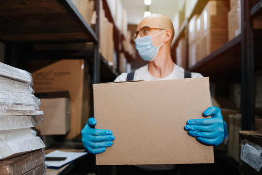 Tired Thoughtul Warehouse Male Worker Carrying Cardboard Box, Standing Between Tall Rows Of Shelves. He Is Wearing Blue Mask And Gloves. Selective Focus On Hands. Bold Head Turned To The Side.