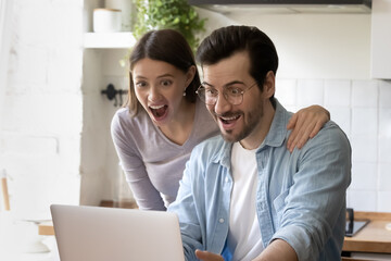Close up amazed young couple looking at laptop screen, sitting at table in kitchen at home, overjoyed family received unbelievable great news, mortgage or loan approval, online lottery win