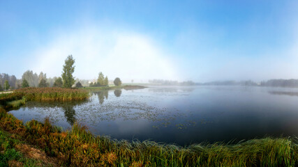 Fototapeta premium rare landscape with a rainbow of fog in the early autumn morning, lake shore, traditional lake meadow vegetation in the foreground, autumn colors in nature, Lielais Ansis, Rubene, Latvia