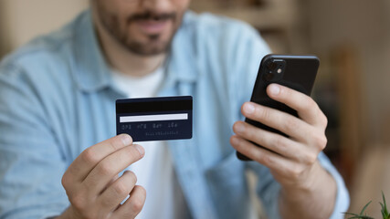 Close up young man holding plastic credit card and phone in hands, making secure internet payment, customer purchasing, shopping online, paying, client browsing banking service, checking balance