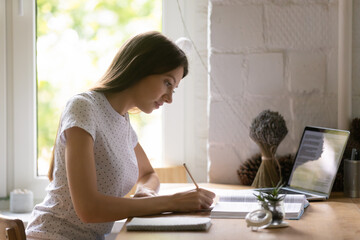 Side view focused woman studying, using laptop, writing notes, concentrated female student working on research project, doing school assignments, reading textbook, sitting at desk at home