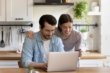 Happy couple using laptop in kitchen at home, making secure internet payment, smiling wife holding plastic credit card, husband browsing online banking service, shopping, purchasing goods
