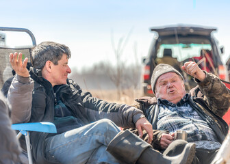Two hunters discuss something emotionally gesticulating, comfortably settled in a forest clearing