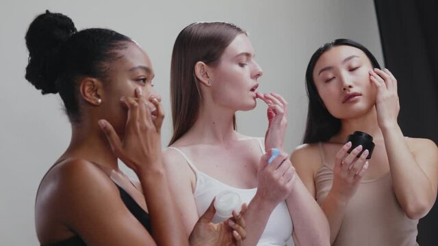 Mixed-race attractive young beautiful women using diverse cream products applying on their faces posing at grey background. Side view. Beauty cosmetics.
