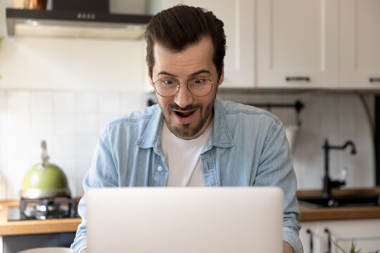 Close up amazed young man wearing glasses using laptop in kitchen, surprised businessman or student with open mouth looking at laptop screen, reading good unexpected news, promotion or lottery win