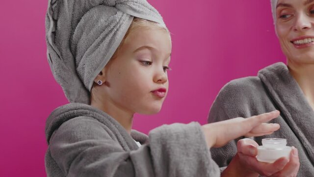 Happy adorable funny girl daughter repeating afte rmom apply face cream looking like beautiful princess. Pink background. Cosmetic ads. Family.