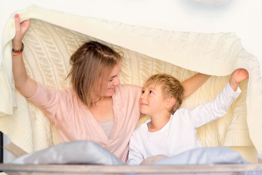 mom and child have fun in the bedroom on the bed, play under the blanket. Young mother with her little son dressed playing in the bed at the weekend together, lazy morning, warm and cozy scene.