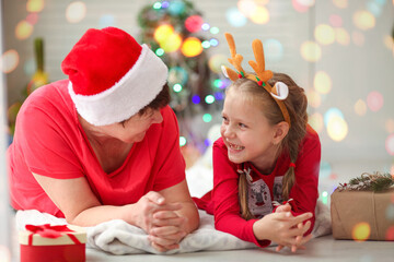 Grandmother and granddaughter look at each other under the Christmas tree and laugh.  Christmas amid the covid-19 pandemic.