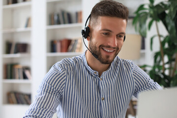 Cheerful young support phone male operator in headset, at workplace while using laptop