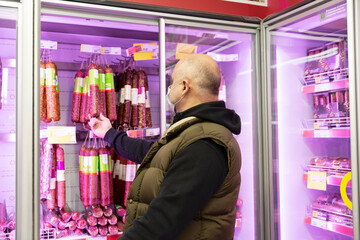 A man wearing a medical mask picks a sausage from a refrigerated display case in a store. Coronavirus pandemic.